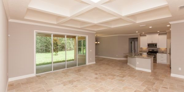 Empty beige living room with sliding glass doors, coffered ceiling, and open kitchen view