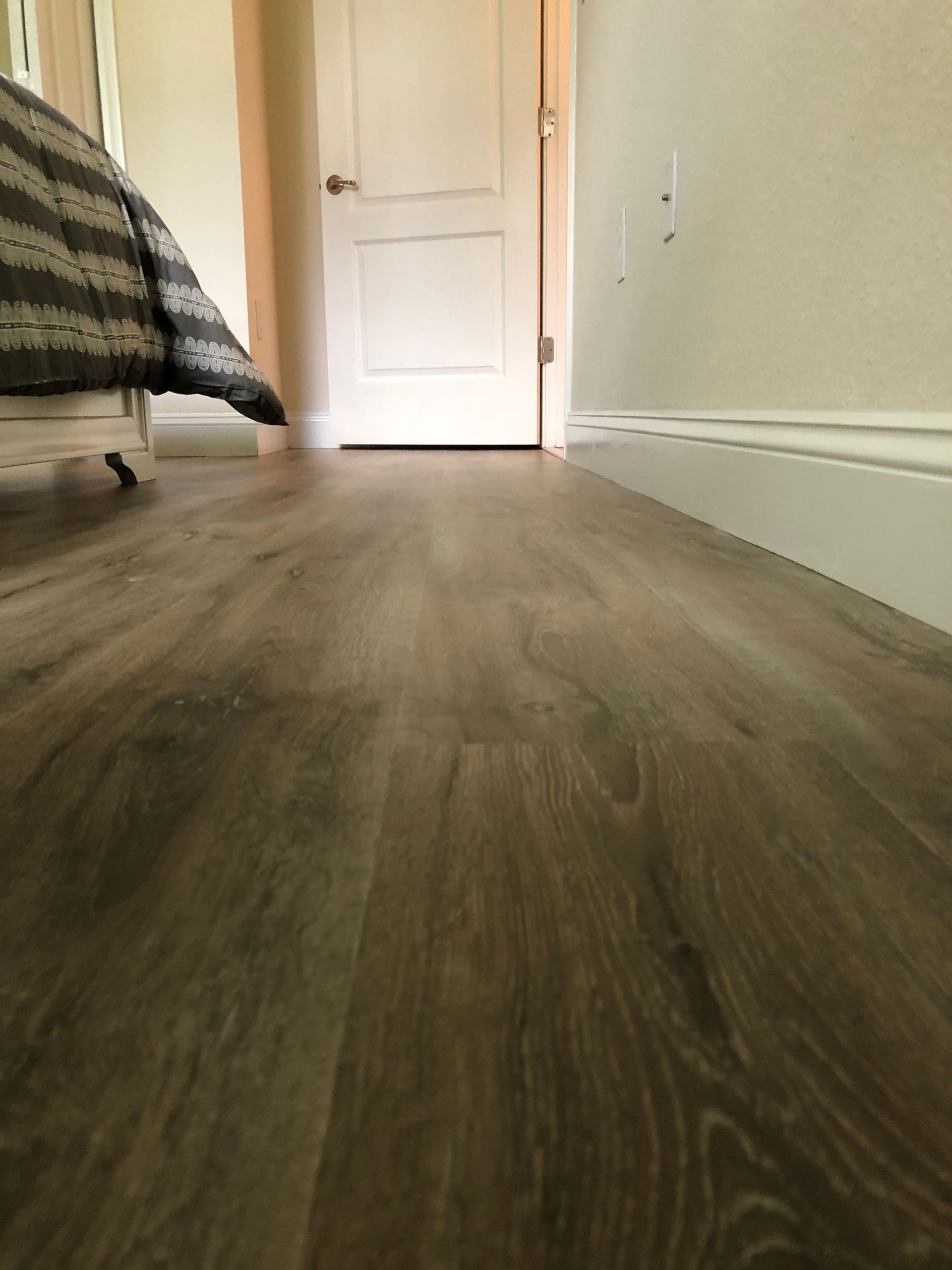 Low-angle view of a hallway with wood flooring, a bed edge on the left, and a closed white door ahead.