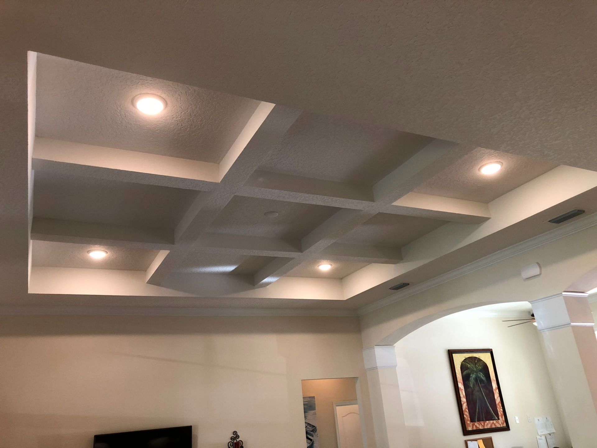 Living room ceiling with recessed tray design, white beams, and warm recessed lights above a wall-mounted TV.