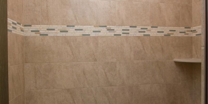 Tan tiled shower wall with a decorative horizontal mosaic border and small corner shelf