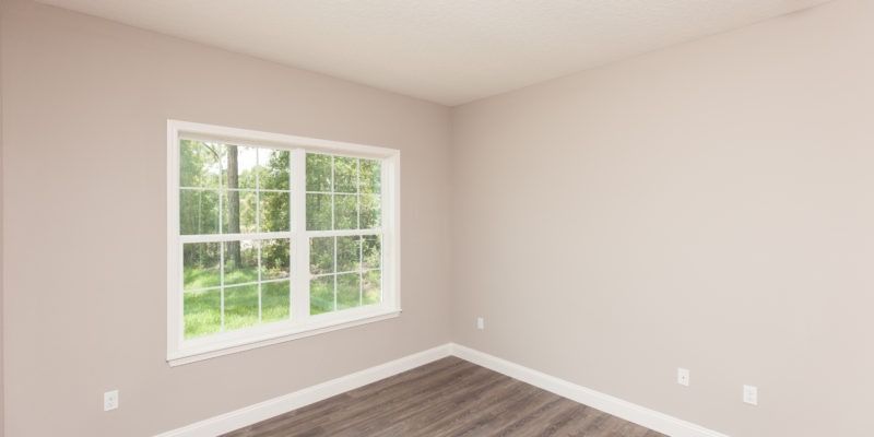 Empty beige bedroom with carpet, white trim, and a large window overlooking greenery