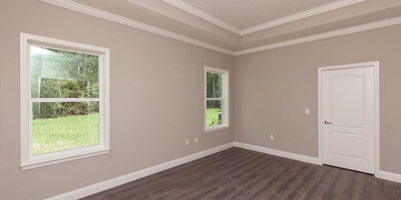 Empty beige room with white trim, two windows, gray wood floor, and a white door