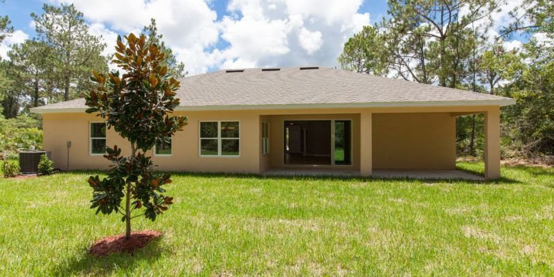 Single-story tan house with a covered porch and a large green lawn under a partly cloudy sky