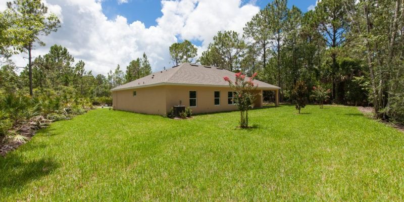 Single-story beige house with a red roof on a grassy lawn, surrounded by trees under a partly cloudy sky