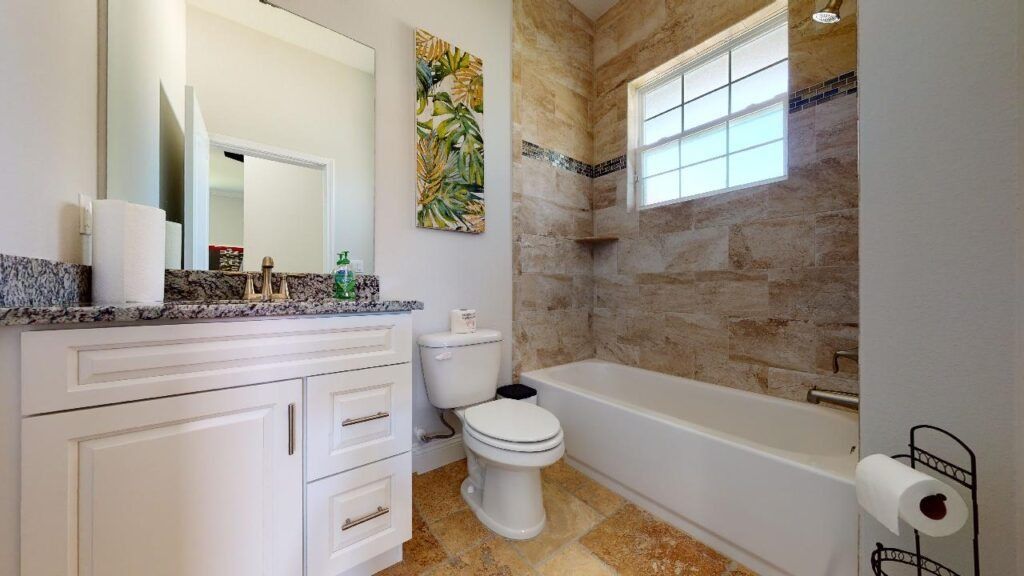 Bright bathroom with white vanity, mirror, toilet, tub, stone tile walls, and a window above the tub