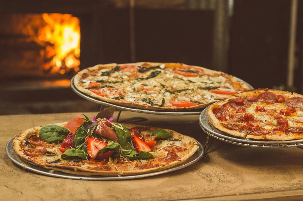 Three pizzas on metal trays, with a wood-fired oven in the background. One pizza has strawberries and spinach.