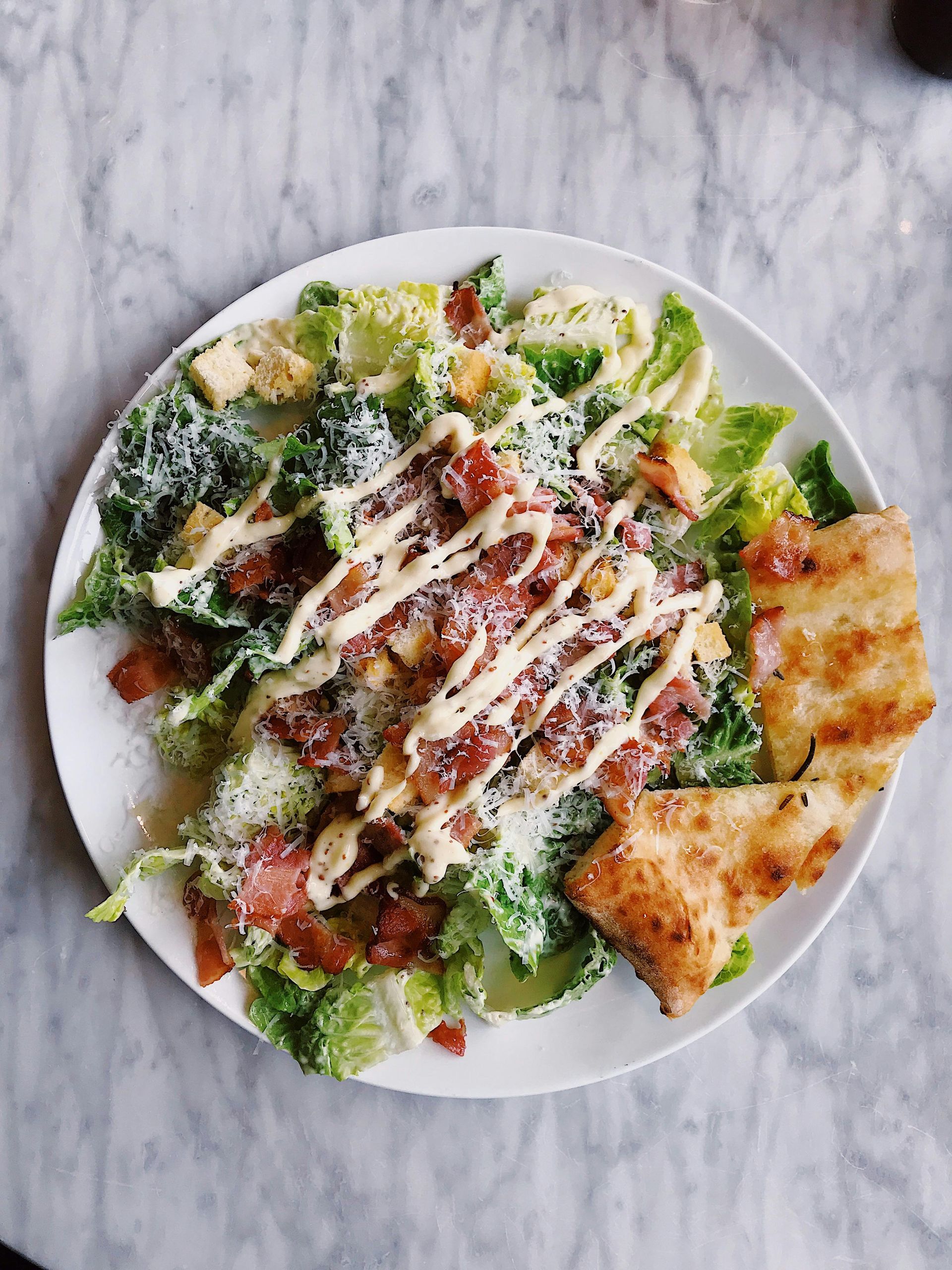 Caesar salad with bacon and parmesan, drizzled with dressing, served with grilled bread on a white plate.