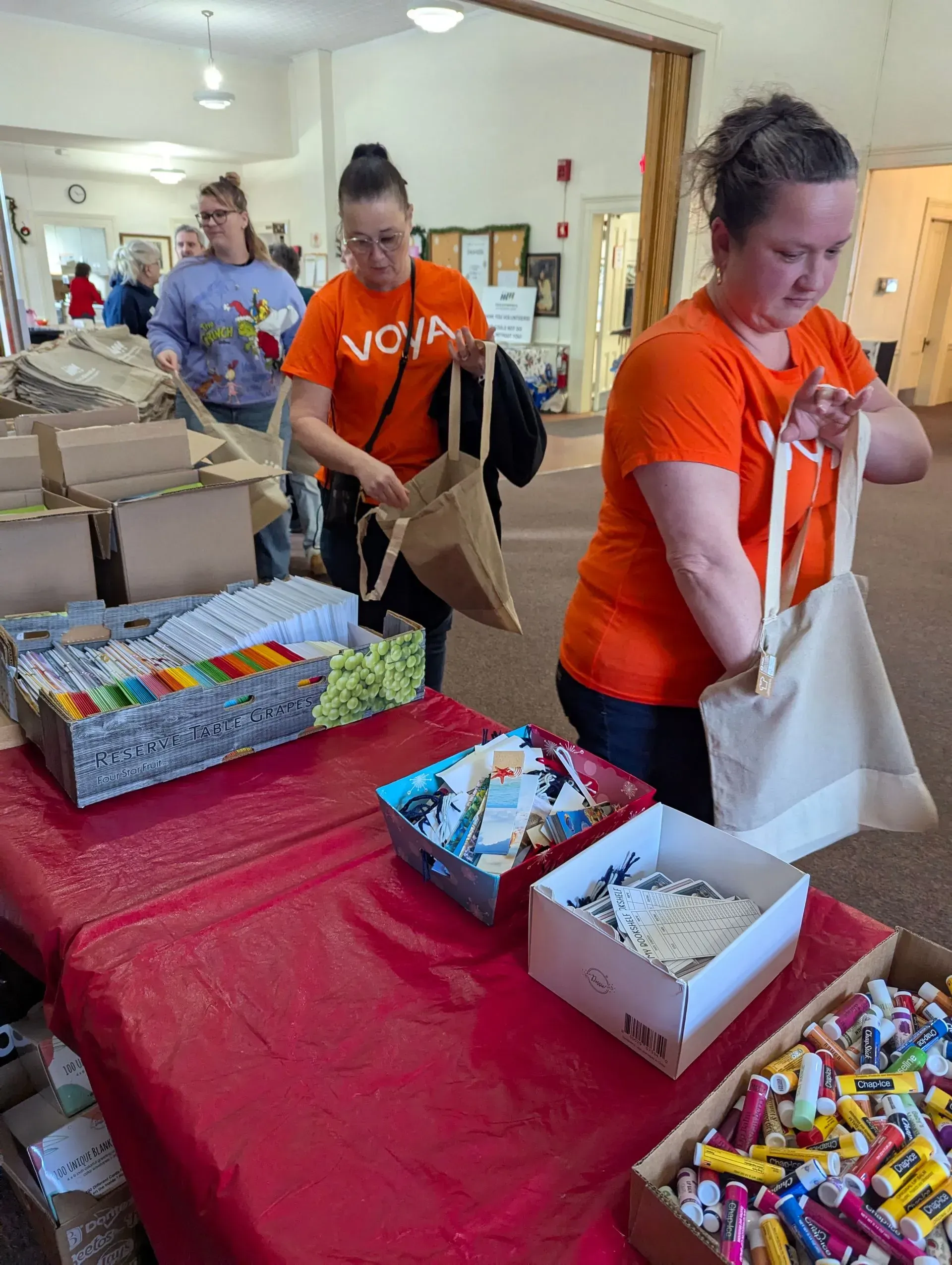 People in orange shirts fill bags at a table with toys and boxes. Interior setting.