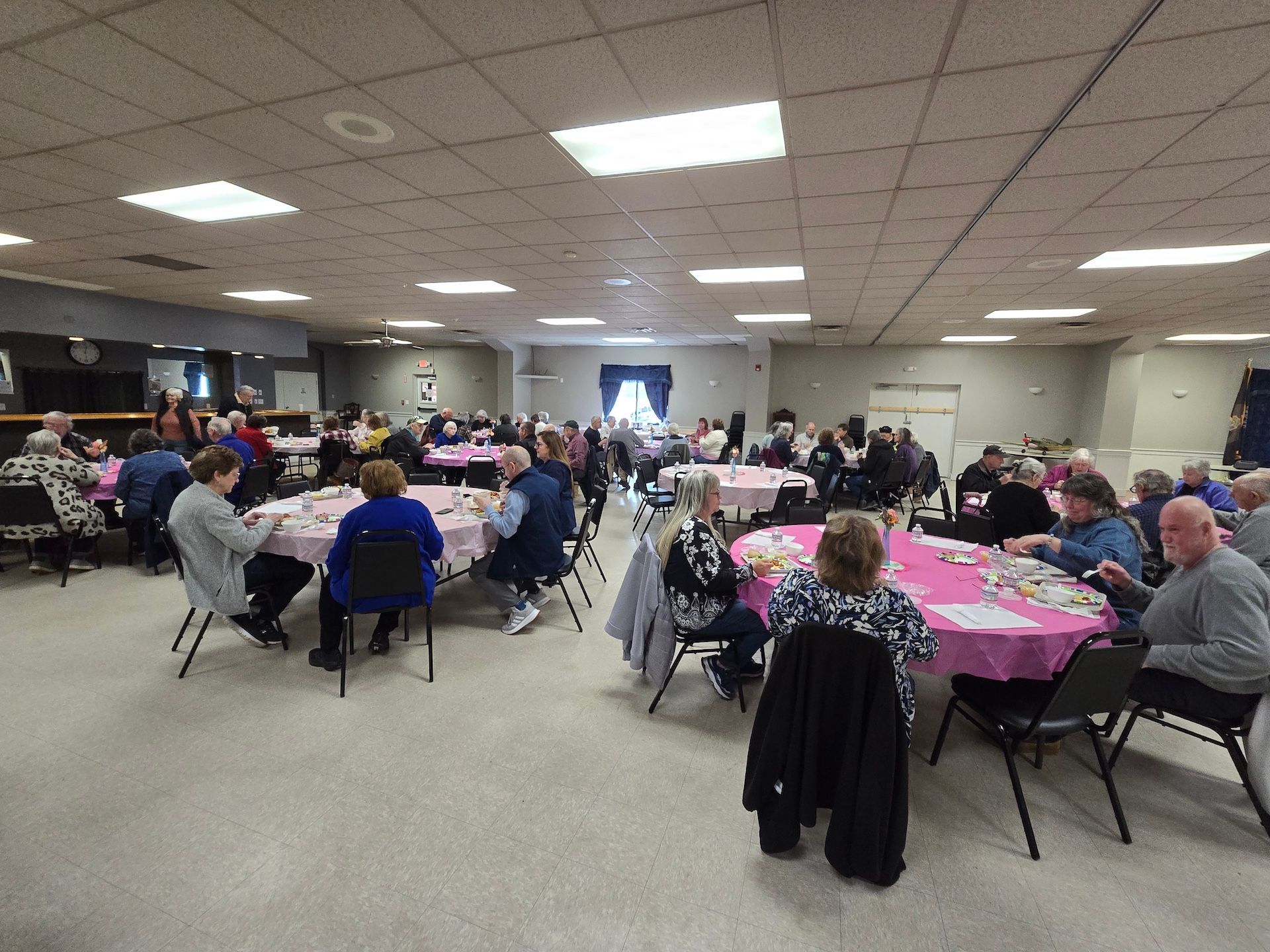 People seated at tables in a brightly lit room, likely a community gathering. Tables have pink tablecloths.