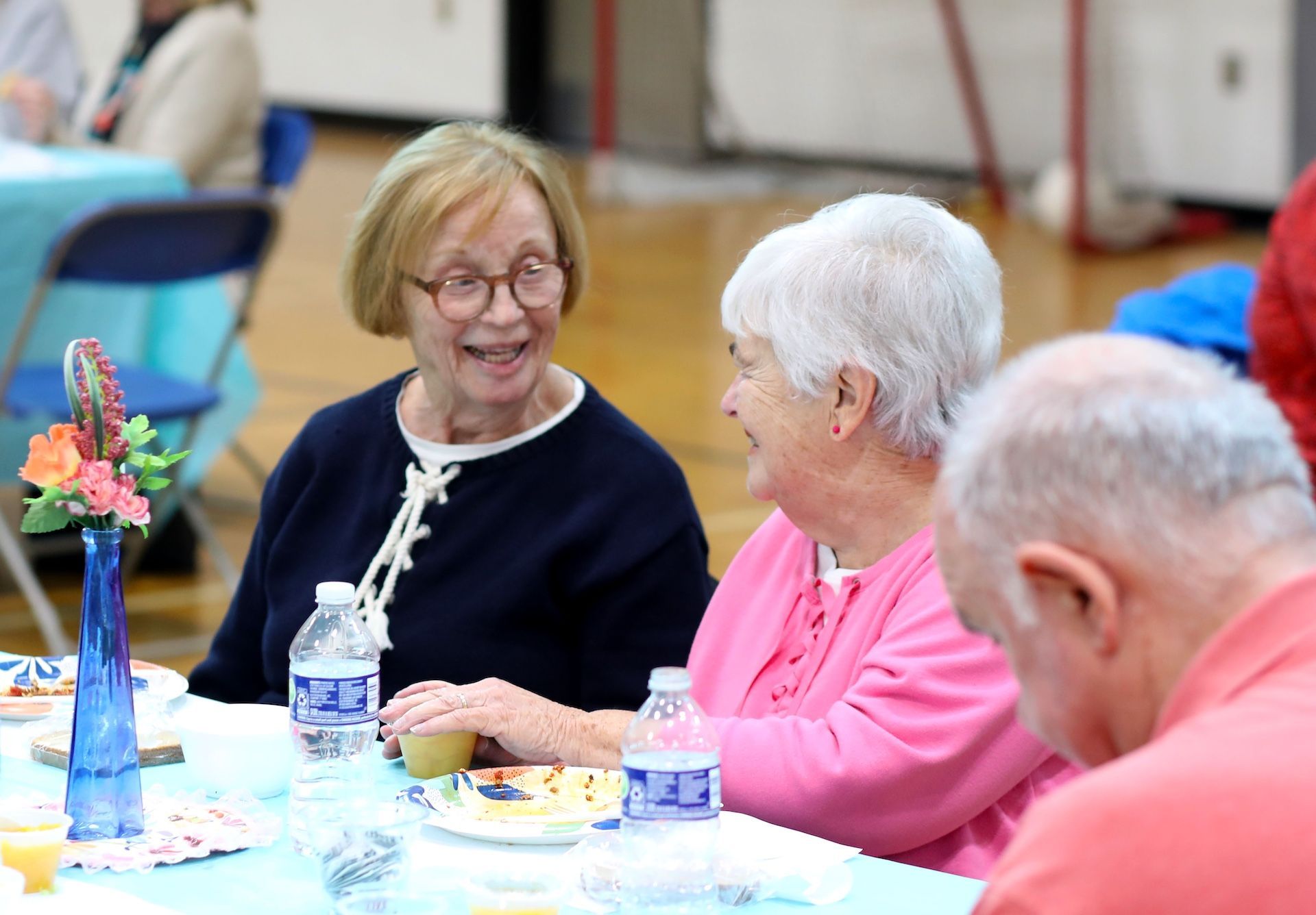 Two older women smiling and talking at a table in a room with a light blue tablecloth and a flower vase.