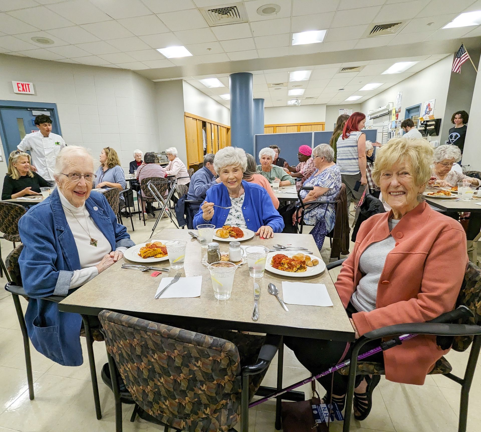 People seated at tables eating in a dining hall. Three women smile at the camera.