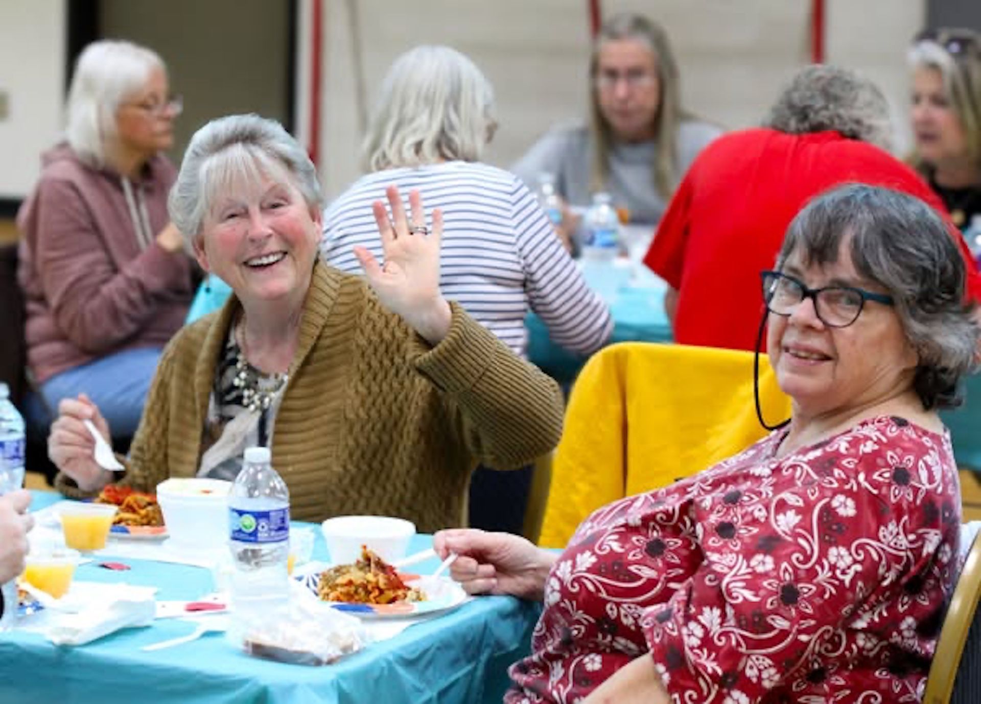 Two women waving and smiling at a table with food, with other people in background.