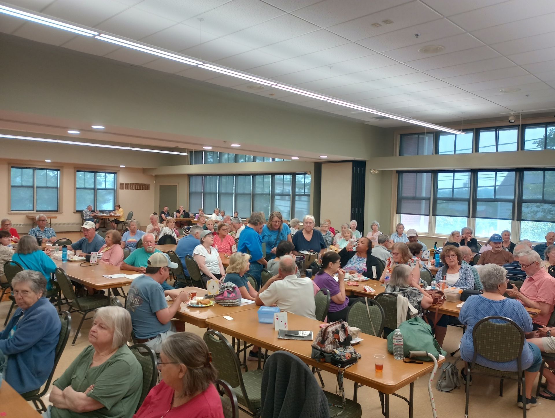 Large room filled with people seated at tables, socializing. Daytime setting, neutral tones.