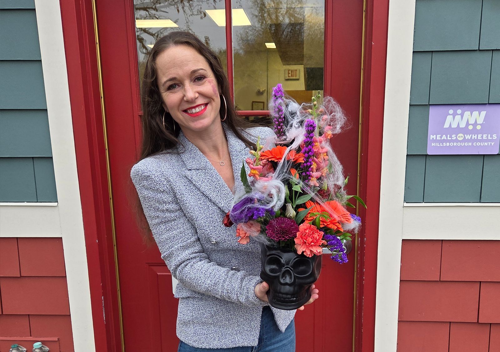 Woman smiles, holding a skull-shaped vase filled with colorful flowers in front of a red door.