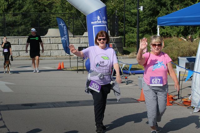Two women wave as they cross a finish line; race. Outdoors, sunny. Woman in pink top, other in purple.