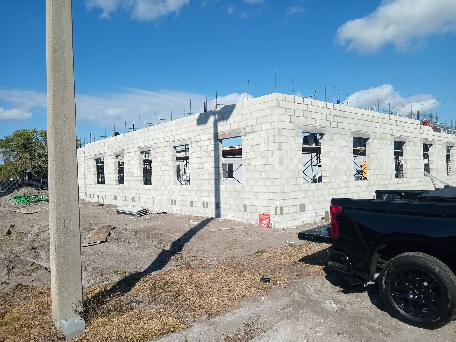 Building under construction, light-colored cinder block walls with window openings, sunny day, black pickup truck parked nearby.