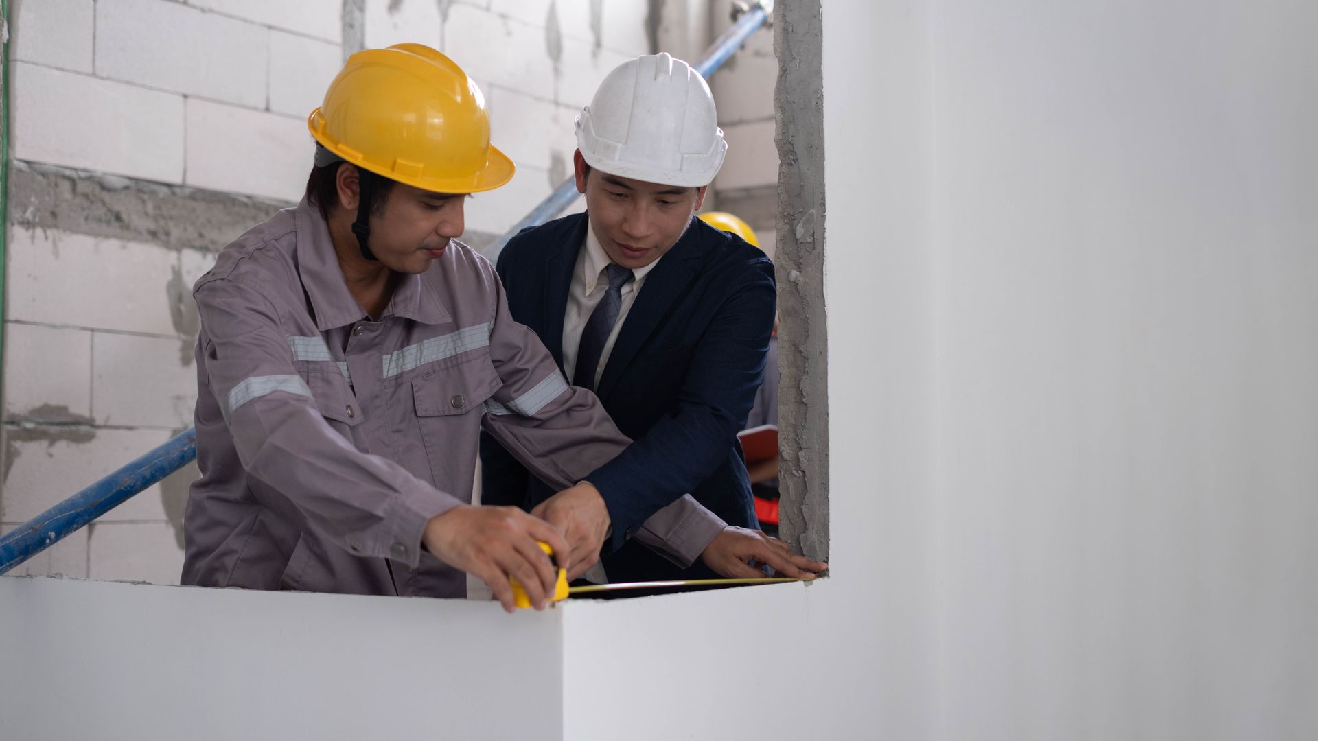 Construction workers and a site supervisor measuring an unfinished interior wall with a tape measure