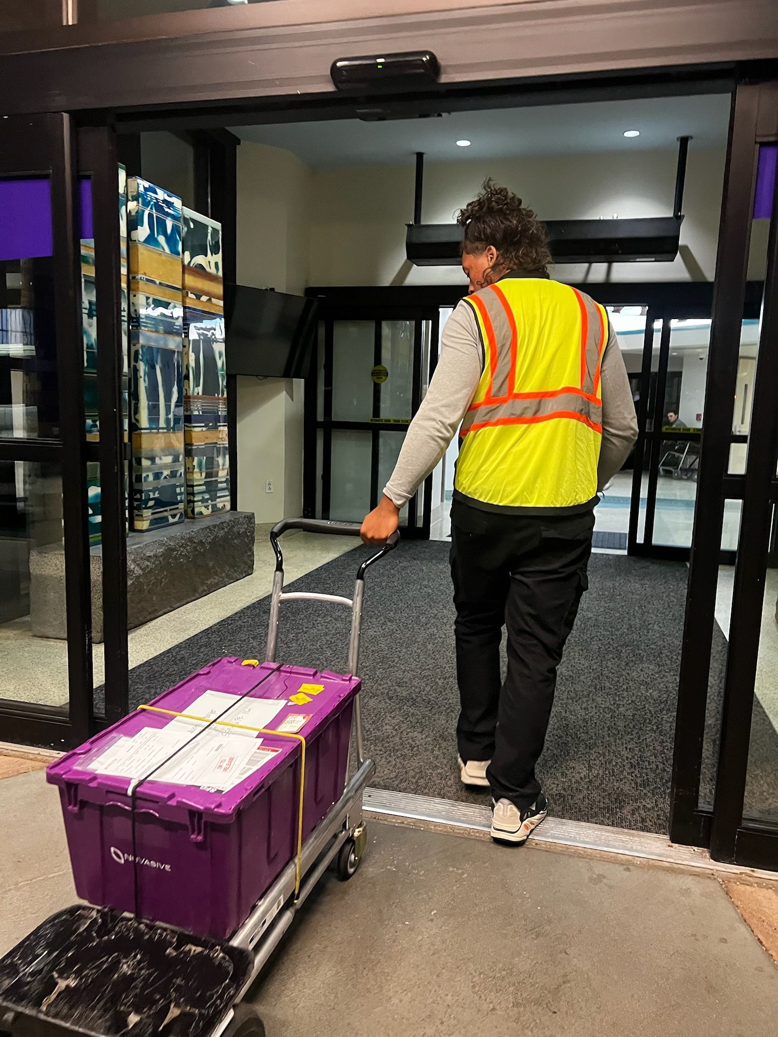 A man in a yellow vest is pushing a cart with a purple box on it.