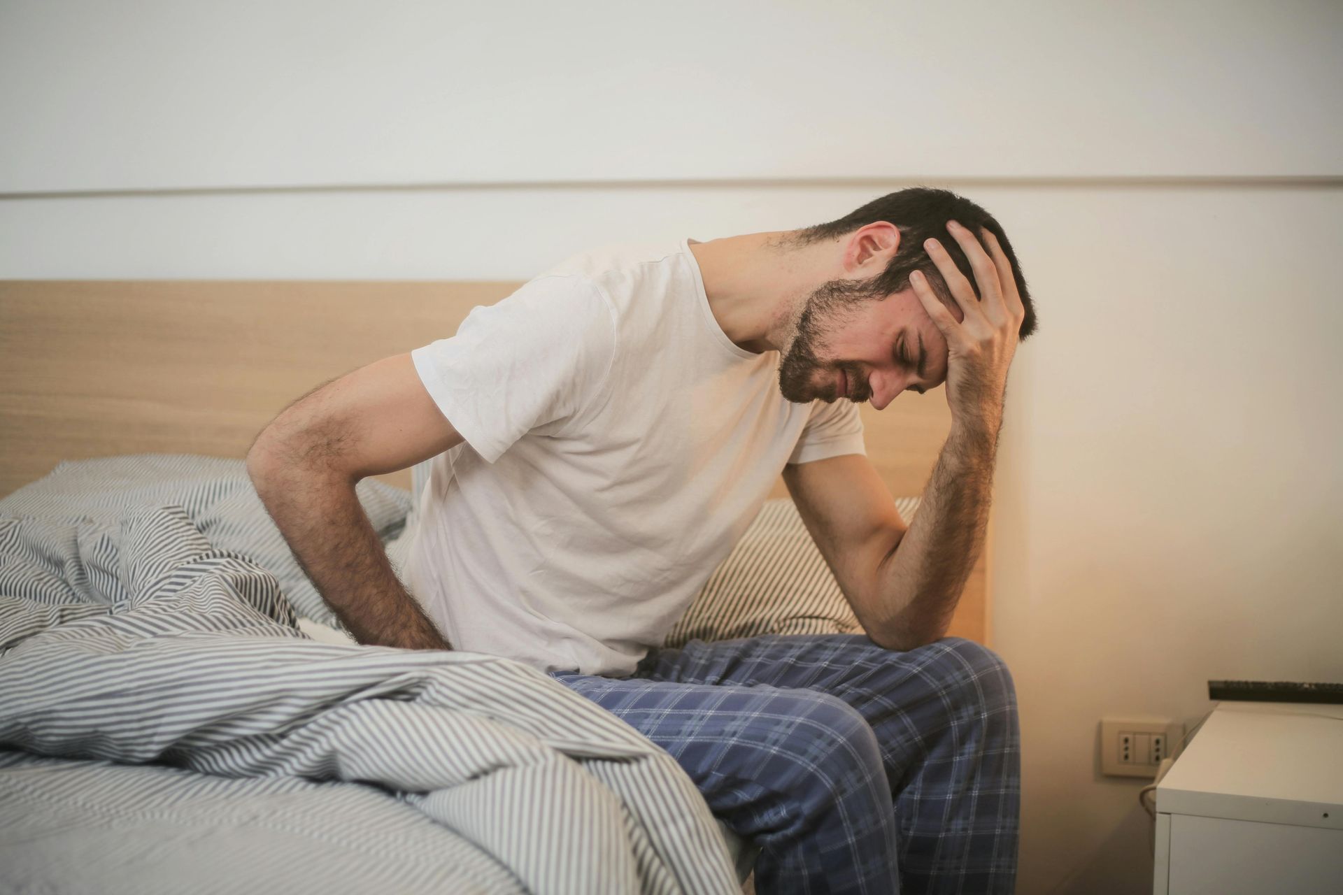 zieke man zittend op de rand van zijn bed in een blauw geruite pyjamabroek en een wit T-shirt met zijn linkerhand op zijn voorhoofd