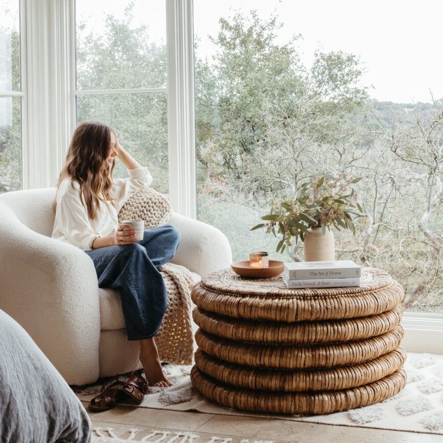 Woman sitting in a white armchair, looking out a large window at a tree-filled landscape. She is filled with thoughts of anxiety and burnout.