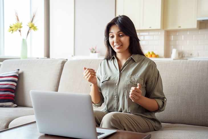 Woman in olive green shirt sits on couch, gesturing while looking at laptop. She is working through stress and anxiety.