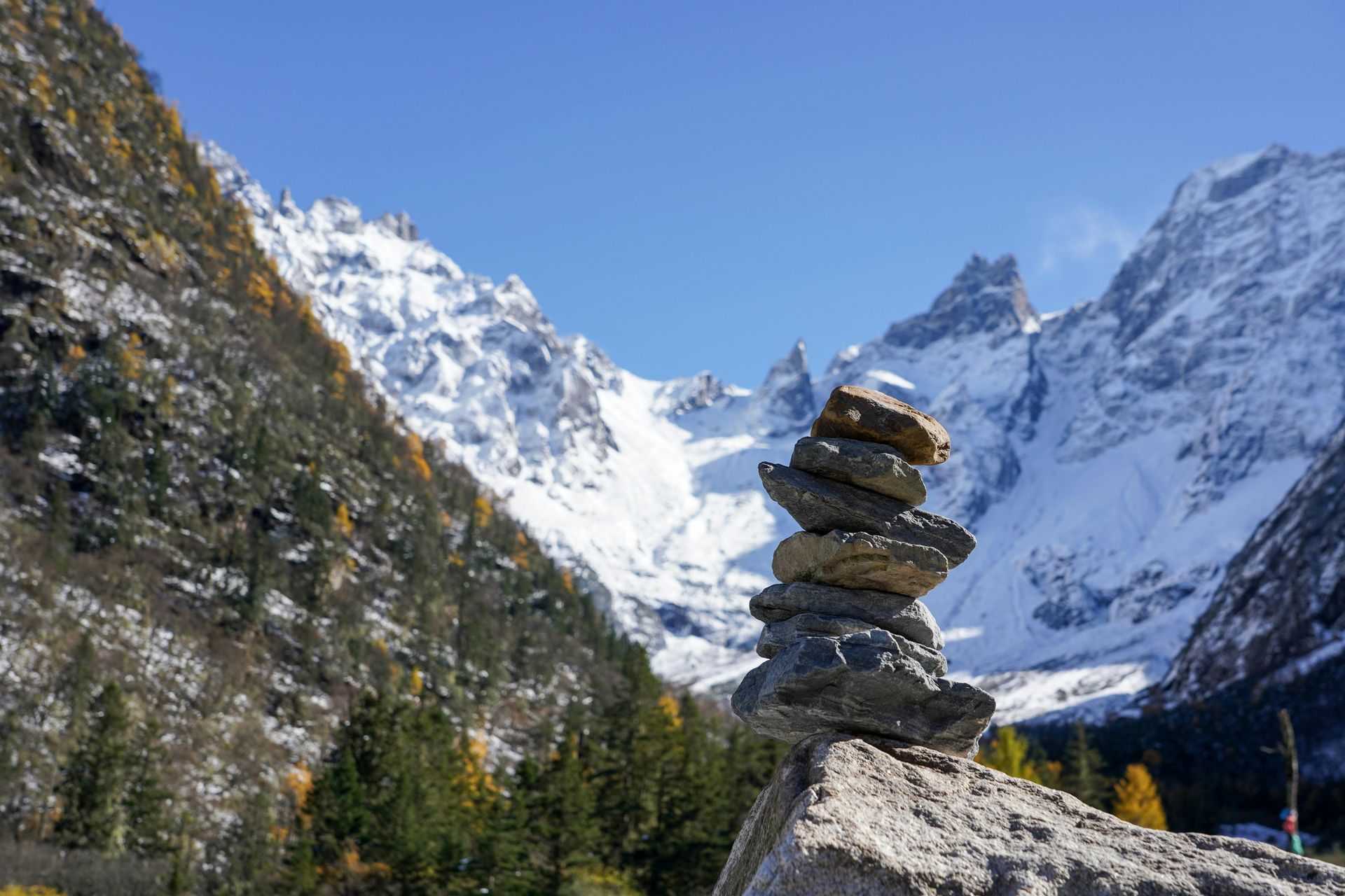 Stack of stones with snow-capped mountains and evergreen trees under a blue sky.