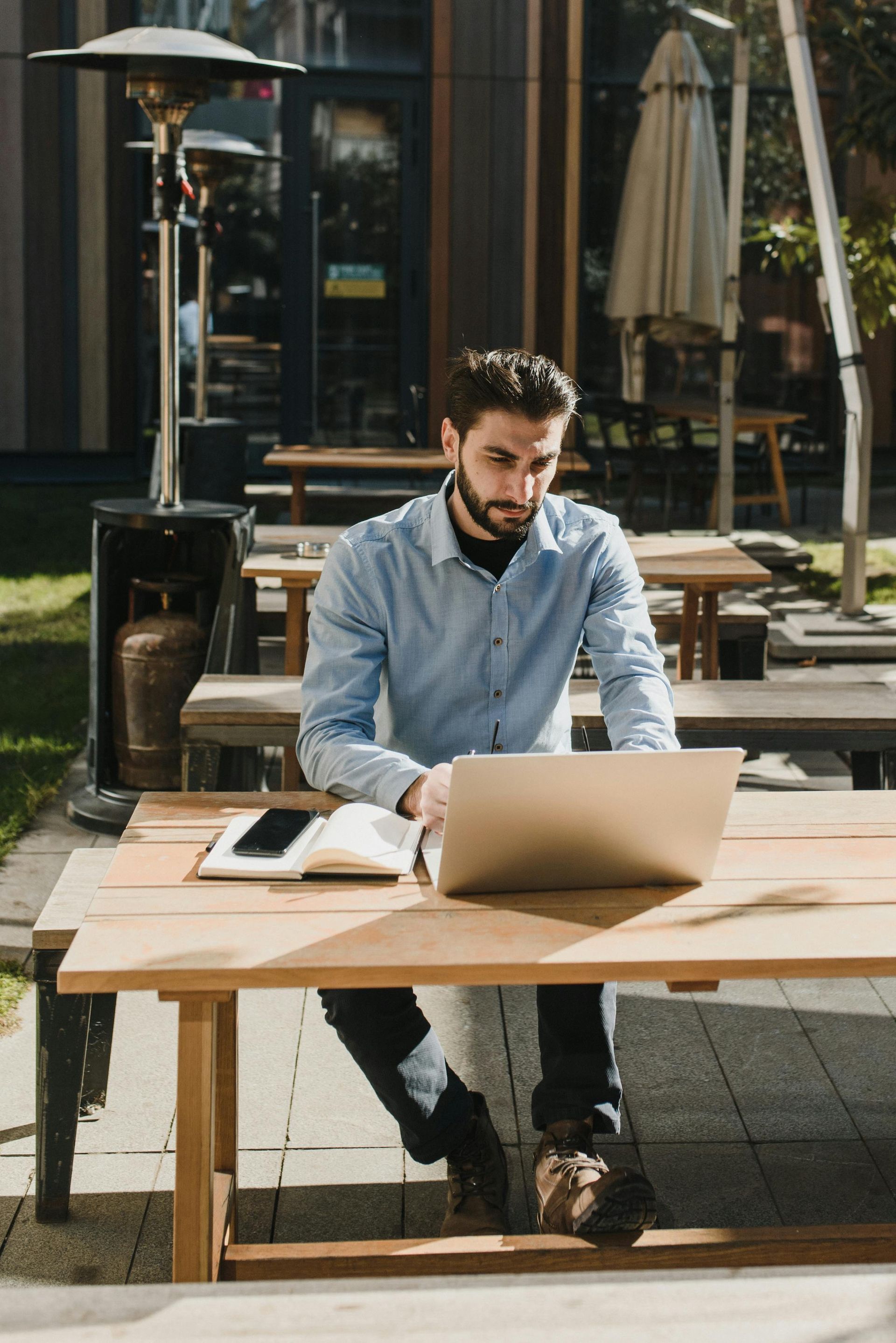 Man at a table with a laptop and coffee, smiling. Indoors, neutral tones, plants in the background.