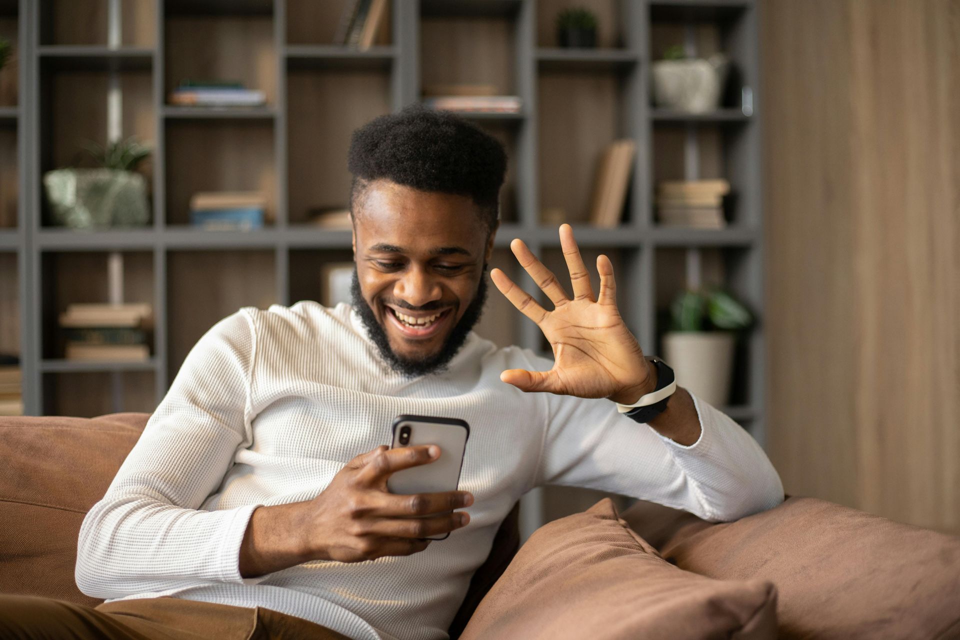 Man on couch waving at phone, smiling. Indoor setting with bookshelves.