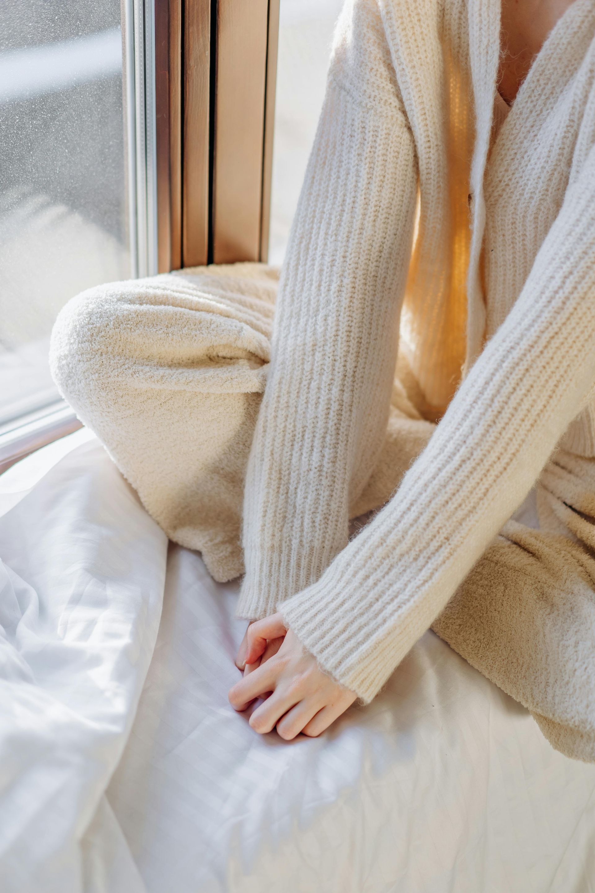 Person in cream-colored clothes sits near a window, hand resting on white fabric contemplating their thoughts.