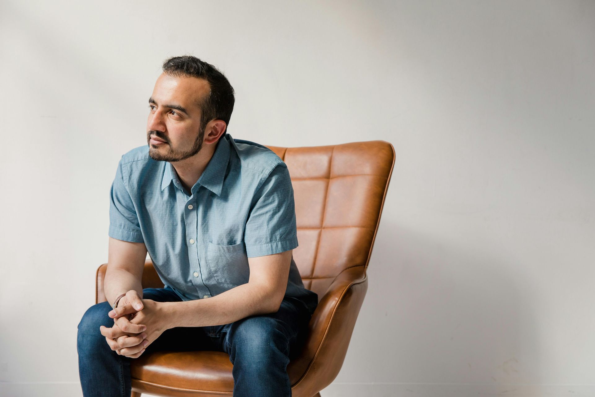 Man sits in a brown leather chair, looking off to the side, hands clasped. Man navigates worry and anxious thoughts.