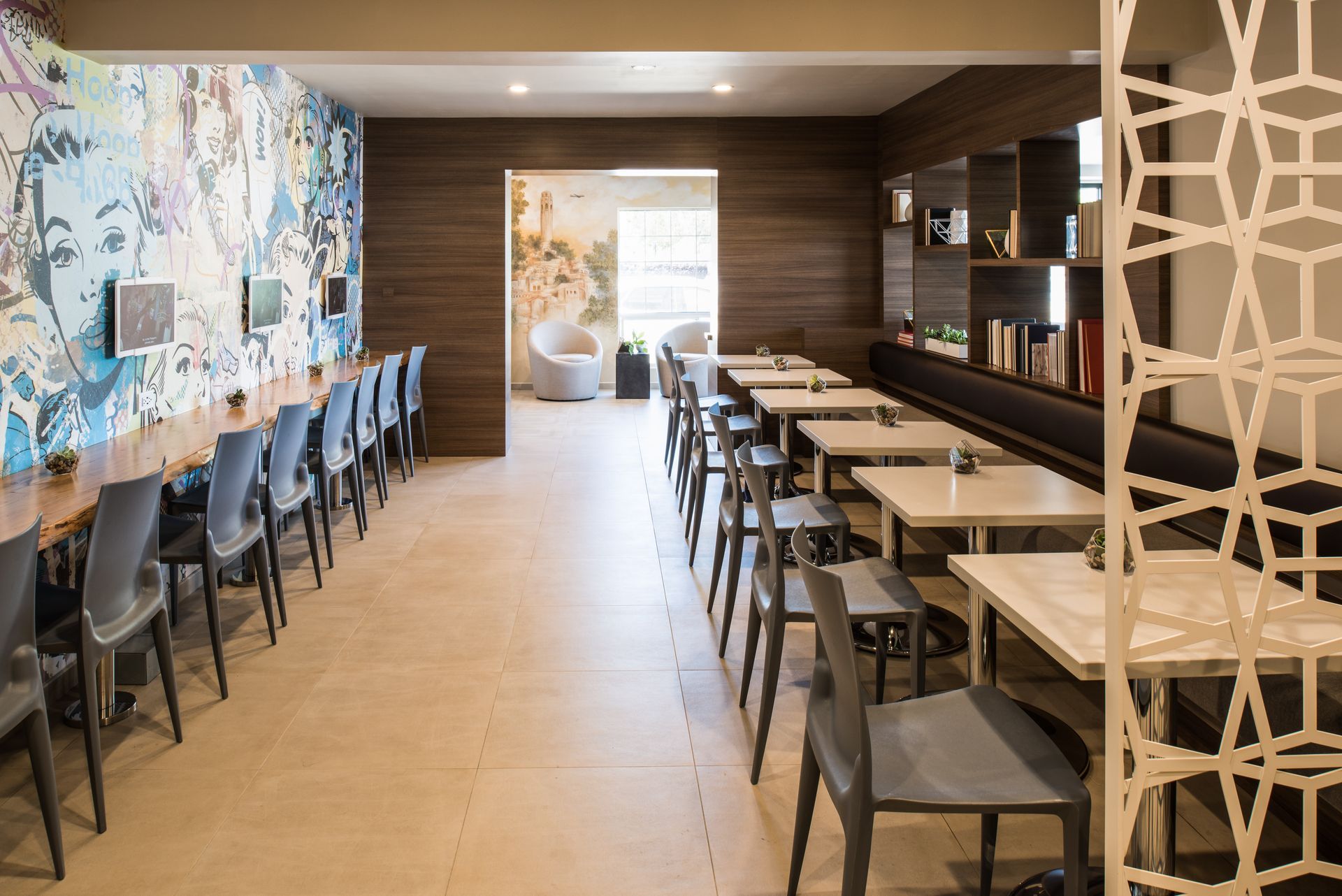 A modern, minimalist cafe or workspace with a wall mural, long bar seating on the left, and square tables on the right.