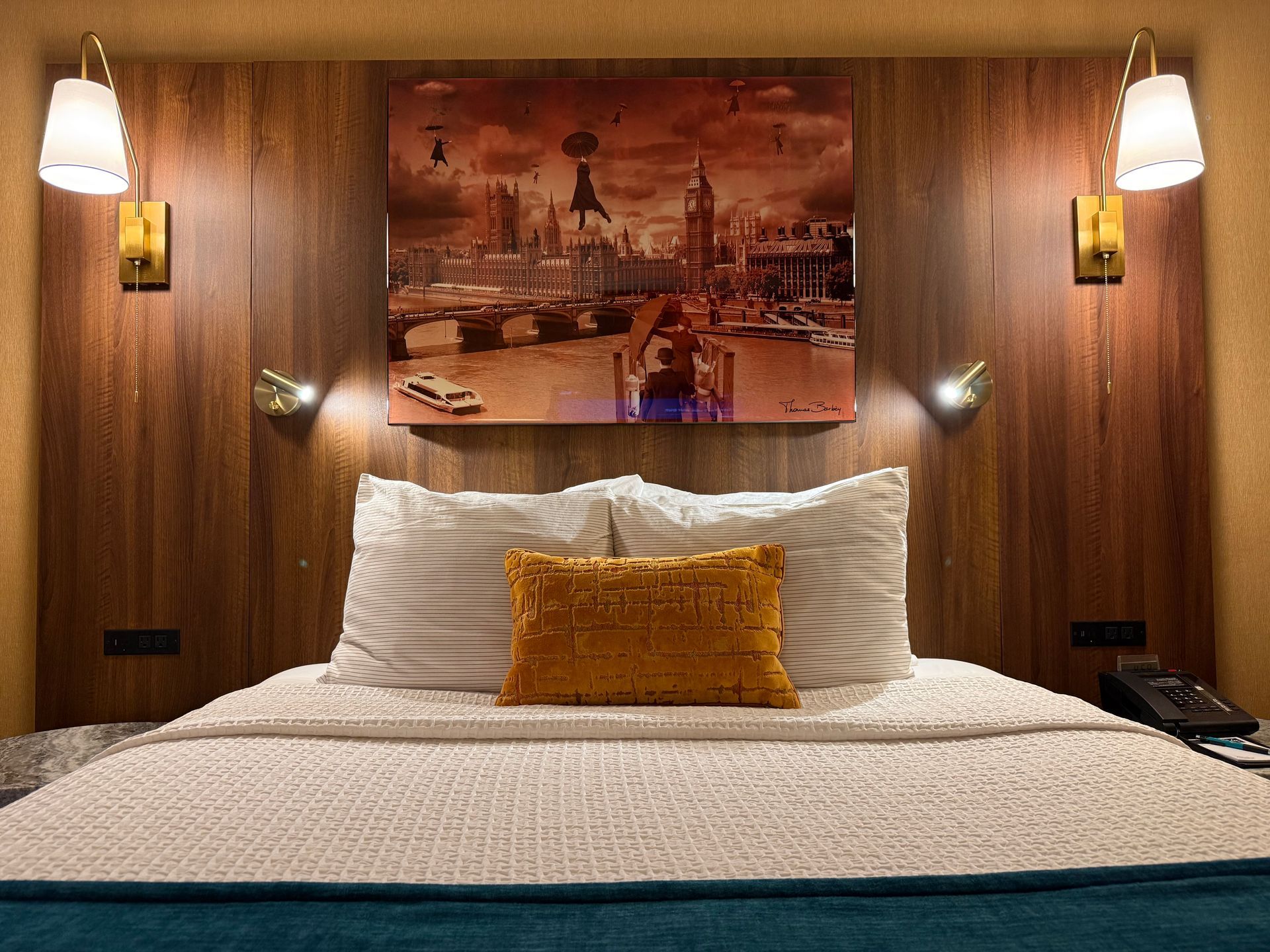 A hotel bed with striped pillows and a mustard yellow accent cushion, set against a wood paneled wall with a London print.