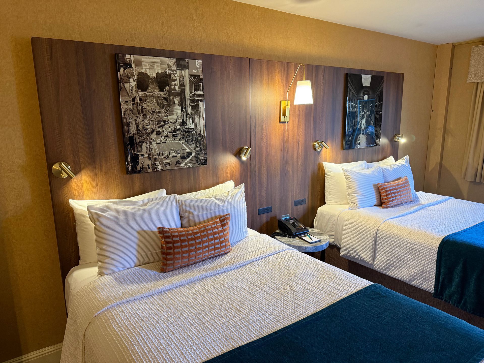 A hotel room with two queen beds, dark wood headboards with wall art, patterned pillows, and white bedding with blue throws.