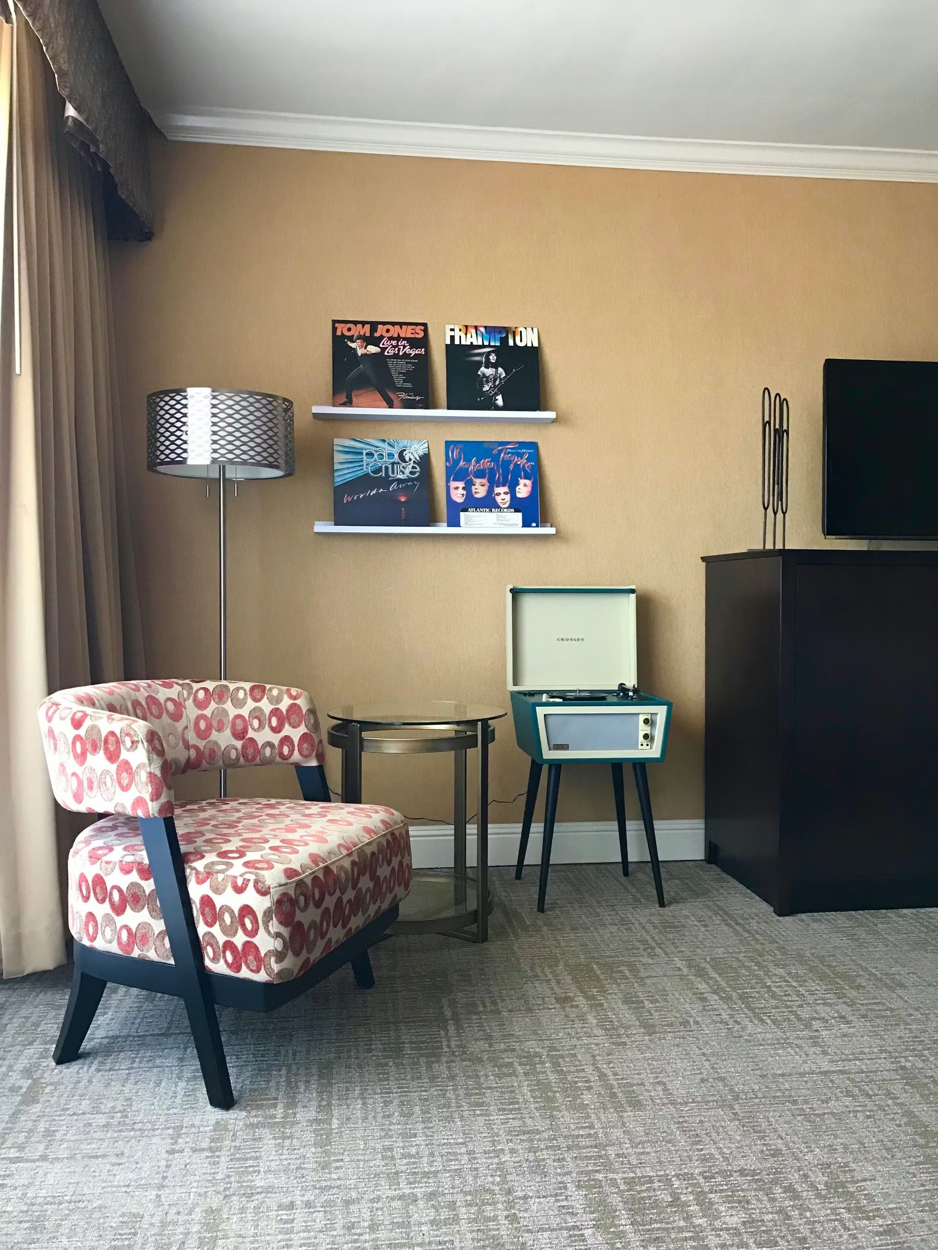 A corner of a room featuring a patterned armchair, a floor lamp, a turntable on a stand, and records displayed on shelves.