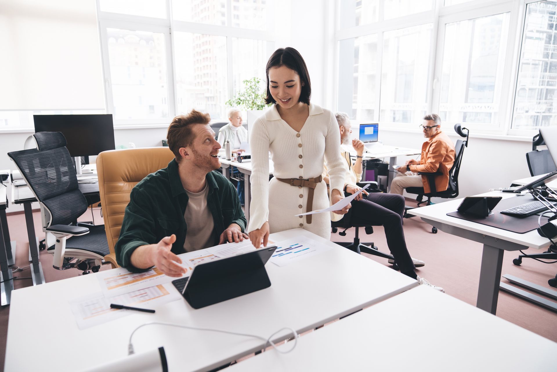 Two coworkers discussing a tablet in a bright modern office, with desks, computers, and office furniture around them.
