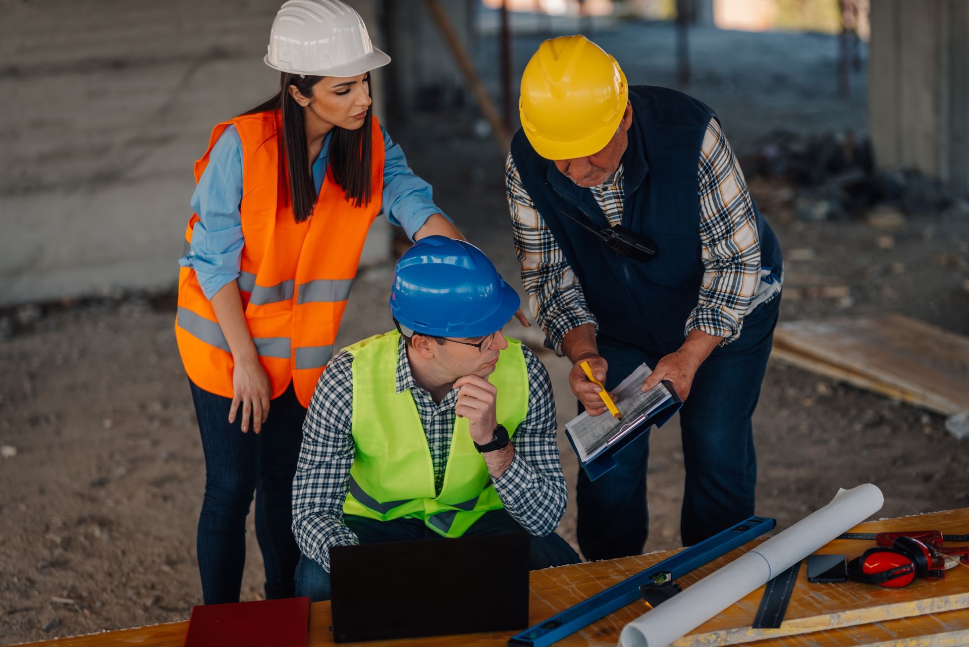 Three construction workers in hard hats reviewing plans at a worksite, one kneeling over a laptop.