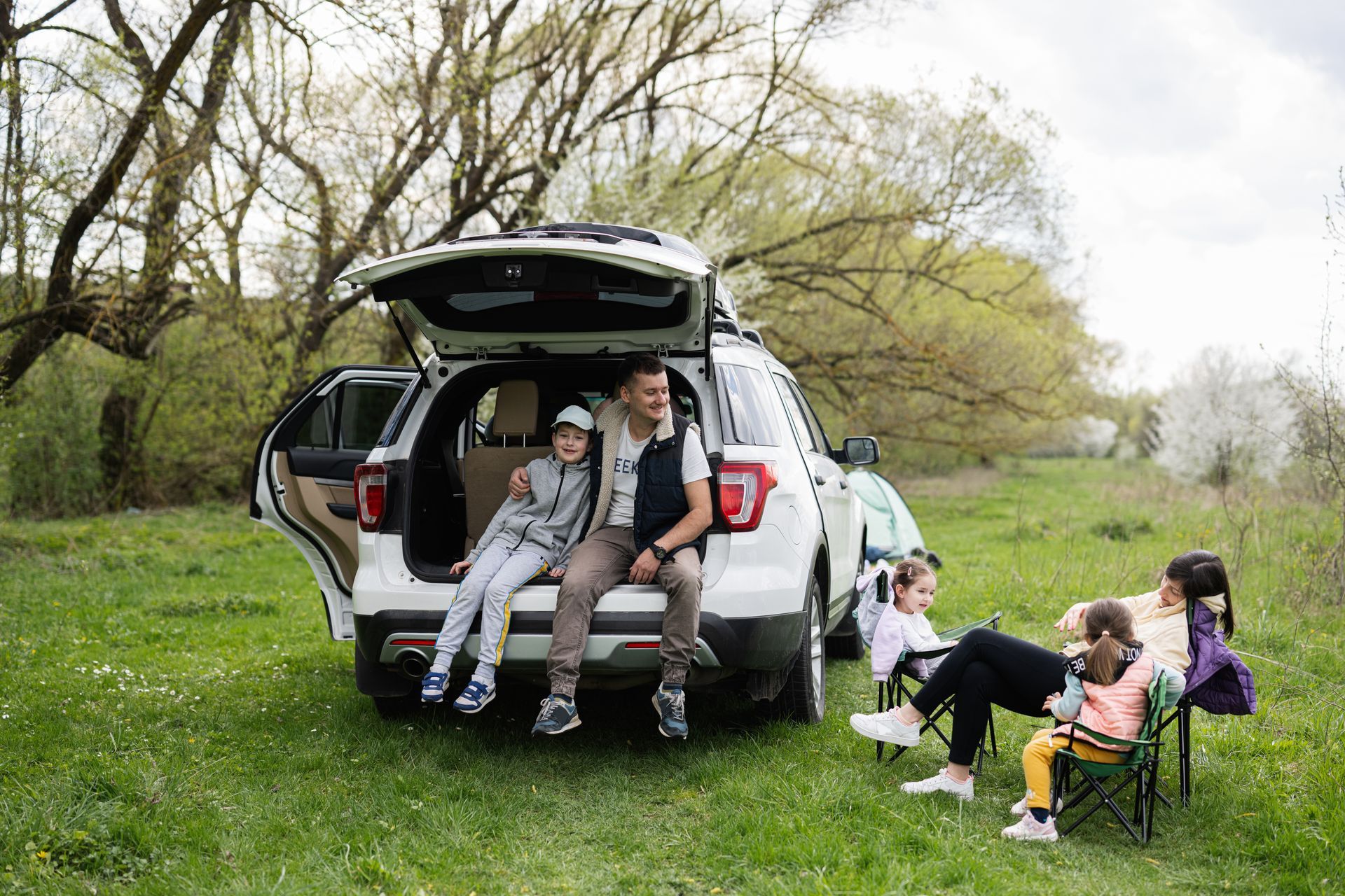 People sitting by an open SUV hatch on a grassy field, with two dogs nearby under trees.