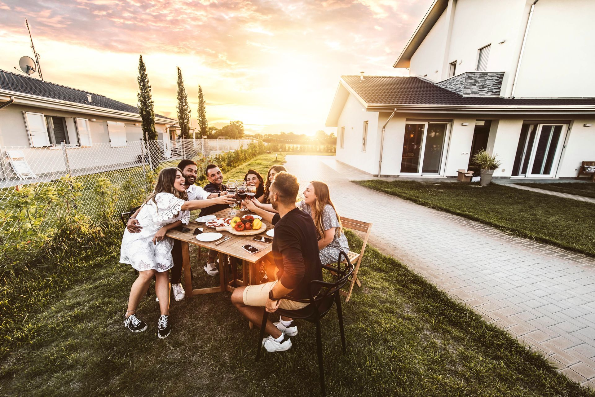 Friends gathered around a table in a sunny backyard between white houses at sunset