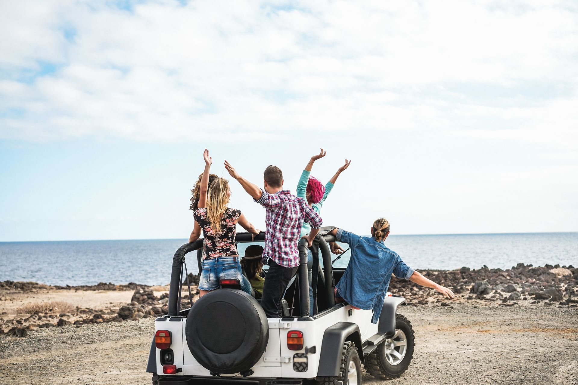 People riding in a white jeep by the ocean, arms raised in celebration.