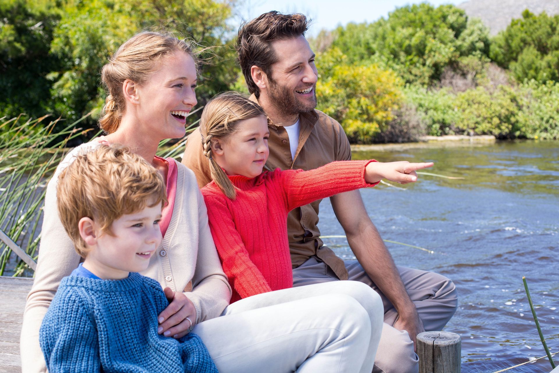 Family sitting by a river, smiling and pointing at the water on a sunny day