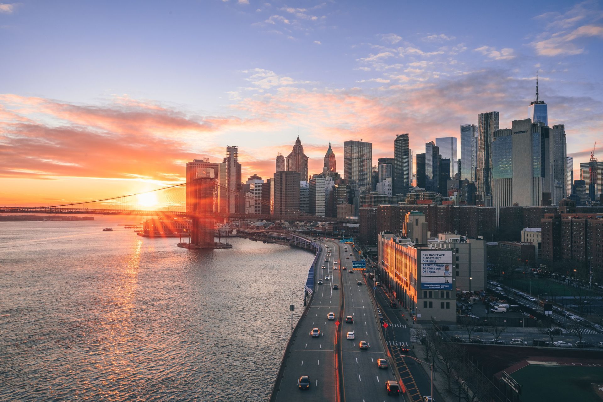Sunset over a coastal city skyline beside a waterfront highway with traffic and calm water