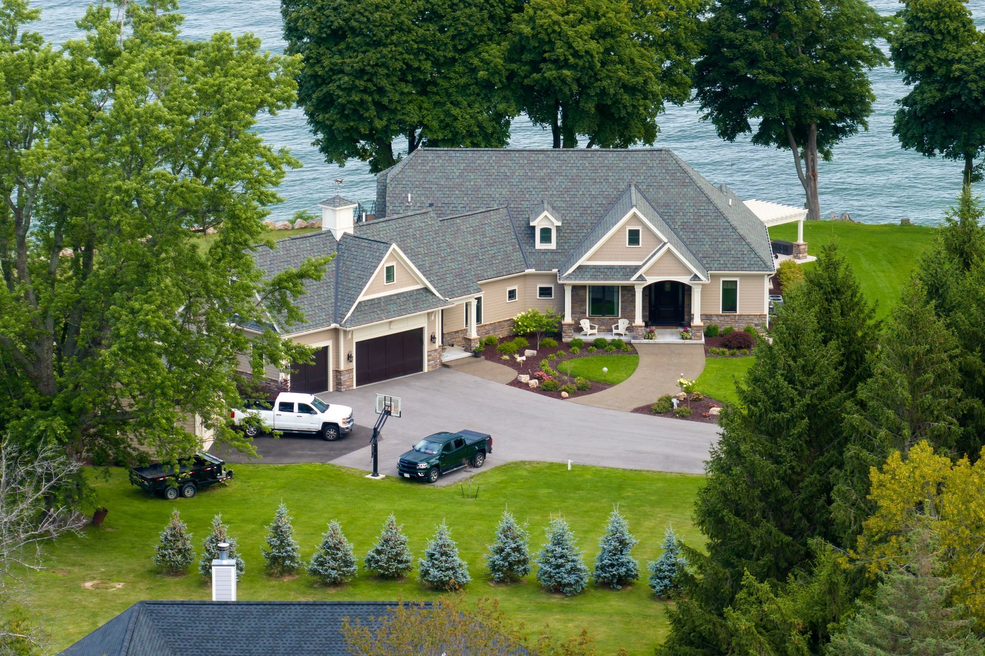 Large tan suburban house with a gray roof, driveway, white car, and landscaped yard surrounded by trees