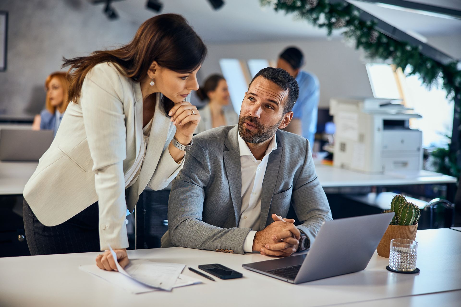 Two coworkers discussing a laptop in a modern office, with documents and a phone on the desk.