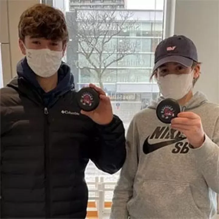 Two young men wearing face masks are holding hockey pucks.