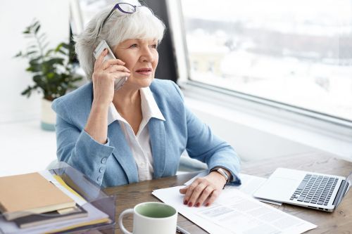 An elderly woman is sitting at a desk talking on a cell phone.
