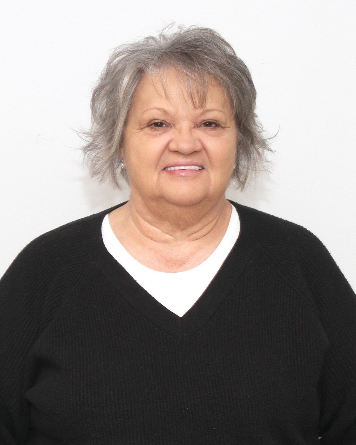 Woman with short gray hair, smiling, wearing a black sweater over a white shirt, against a white backdrop.