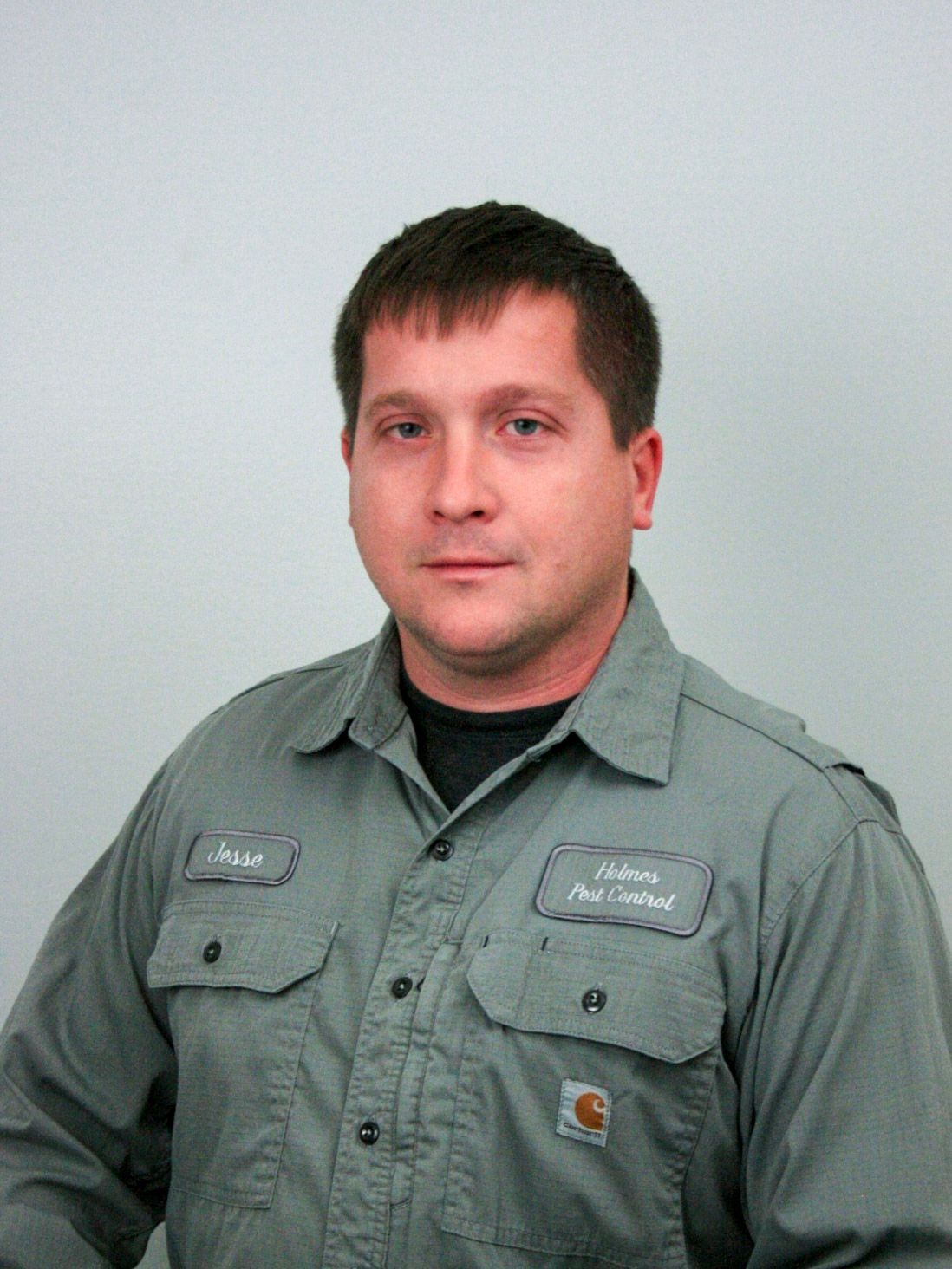 Man in a gray work shirt with name tags, standing in front of a white wall.