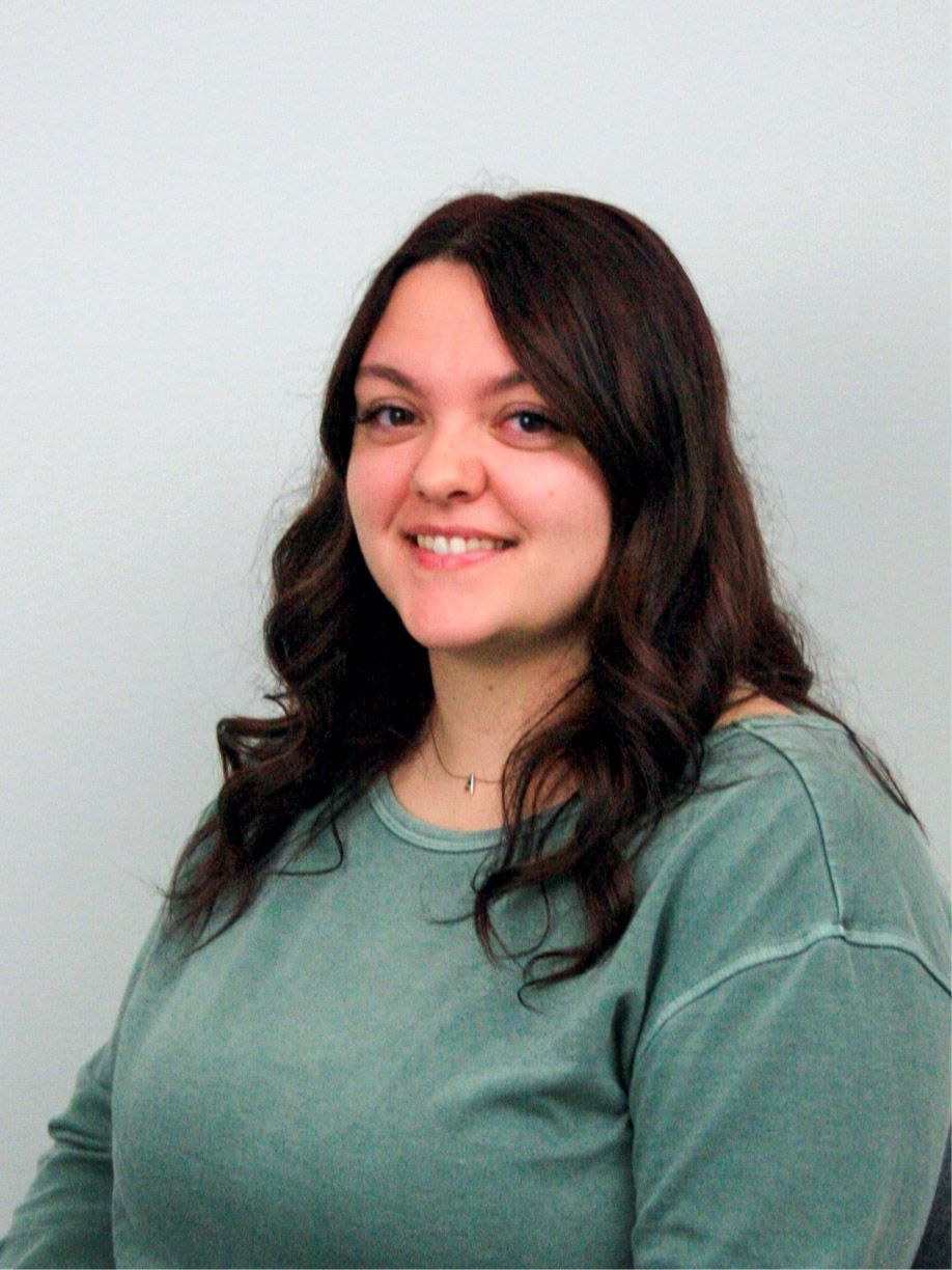 Woman with brown hair smiling, wearing a teal sweater, in front of a white wall.