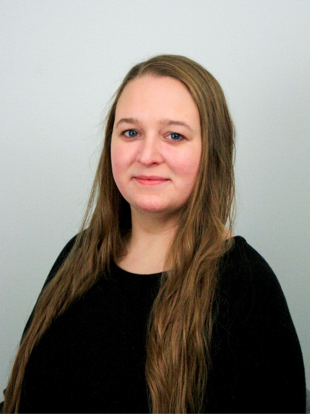 Woman with long brown hair, wearing a black top, smiling, against a light gray wall.
