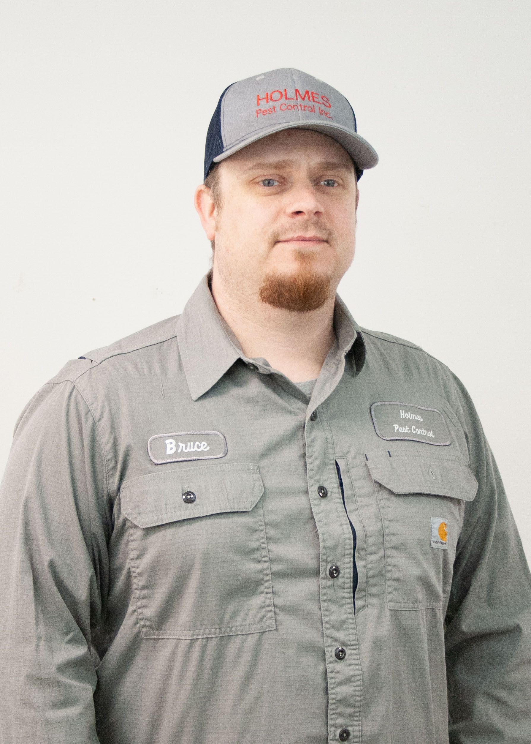 Man wearing a grey work shirt and a baseball cap with company logo. Standing against a white background.